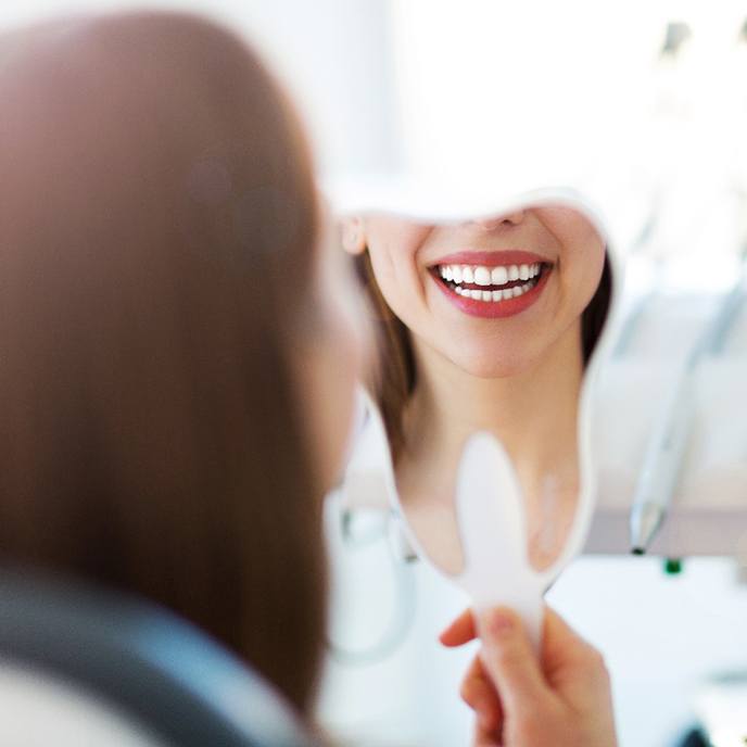 A woman looks at her smile in the mirror while seeing her cosmetic dentist in St. Johns